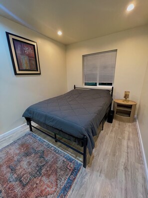 A bedroom with a bed, a nightstand, a lamp, a window with blinds, a framed picture on the wall, and a patterned rug. - Brand New Private ADU (Los Angeles)
