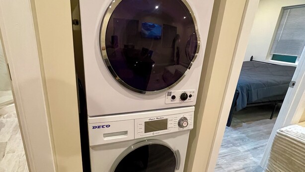 En kompakt tvättmaskin som monteras på väggen, med en digital display och en kontrollpanel, placerad i ett rum med trägolv och en säng i bakgrunden.