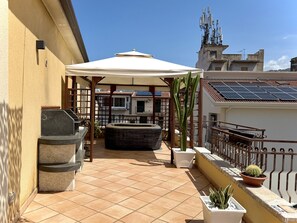 Un patio al aire libre iluminado por el sol, con una parrilla, un parasol blanco y macetas con plantas.