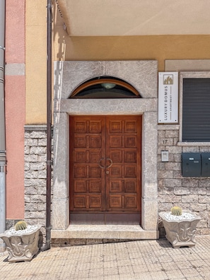 Une porte en bois à deux battants, surmontée d’un arc en pierre, une enseigne indiquant qu’il s’agit d’un appartement de luxe, et deux plantes en pot de chaque côté de l’entrée.