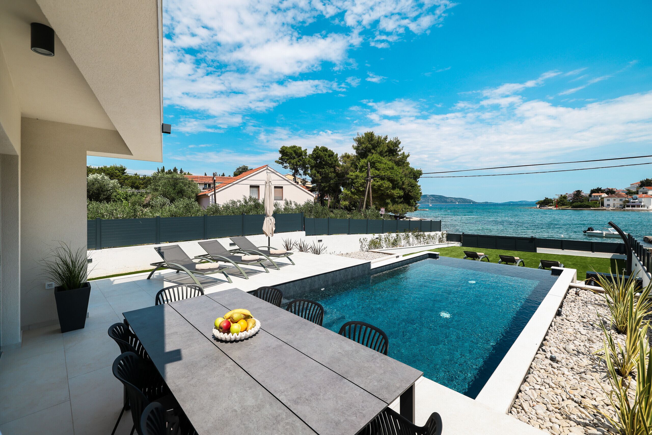 A swimming pool with a table and chairs, a view of the sea, and a building in the background.