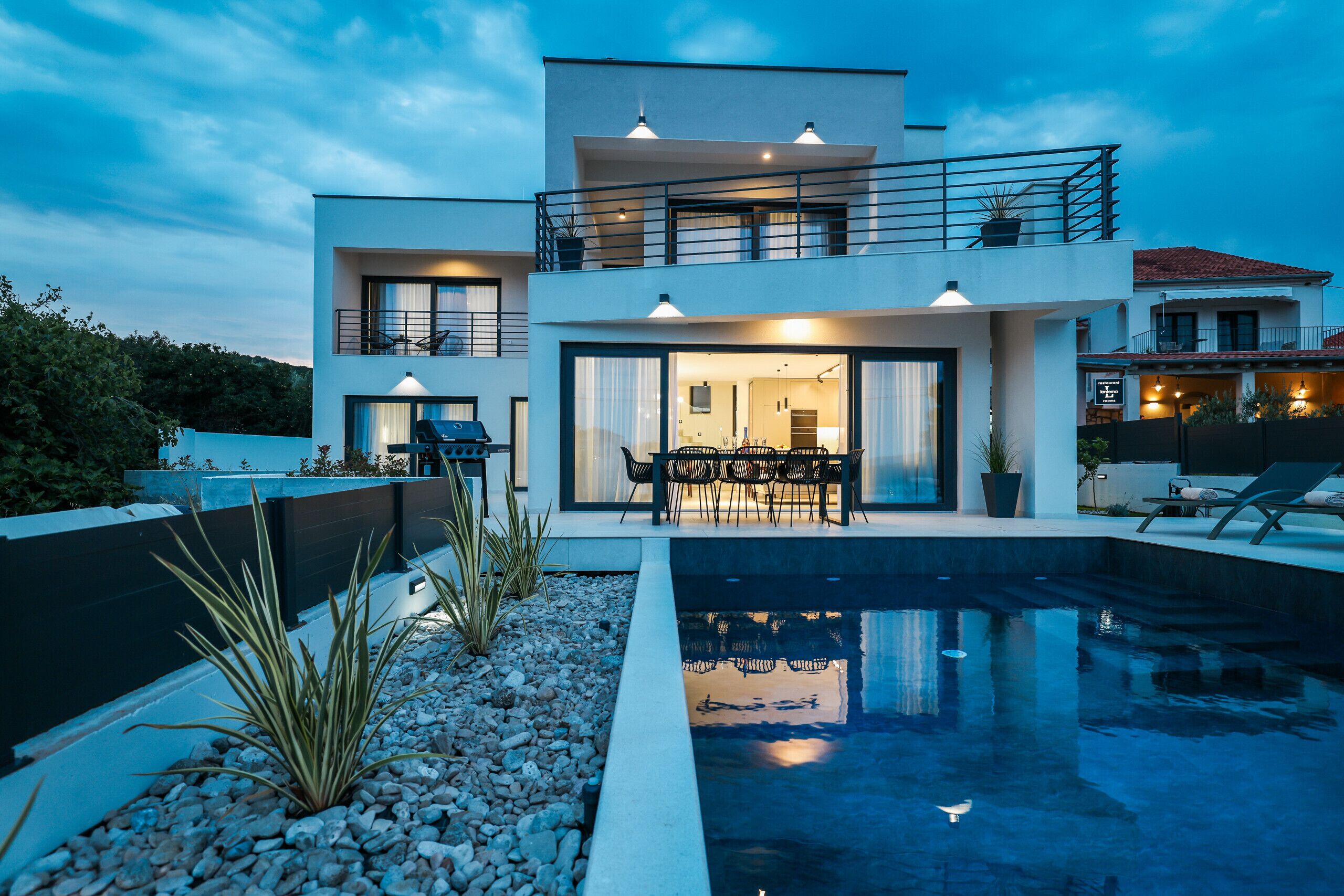 Une maison moderne à deux étages dotée d’une piscine, d’un espace extérieur aménagé et d’un coin repas.