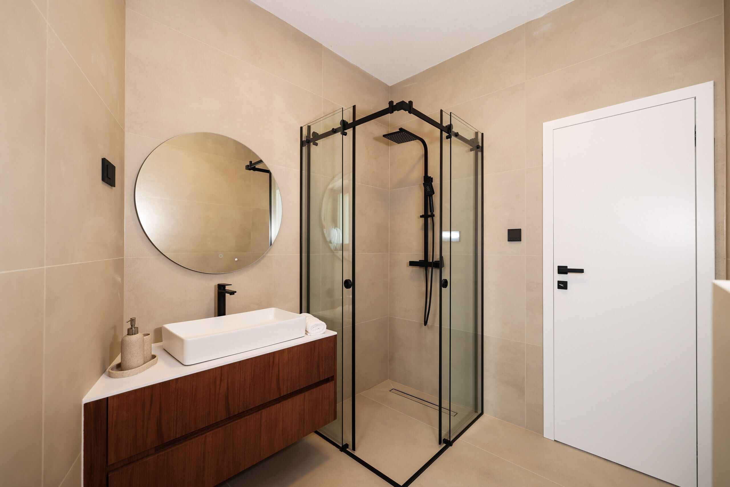 A modern bathroom with a glass-enclosed shower, a round mirror, and a wooden vanity.