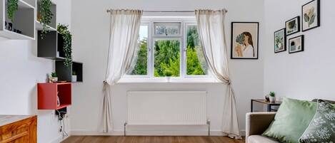 Una sala de estar con ventana, radiador, sofá, mesa de centro y estanterías con plantas.