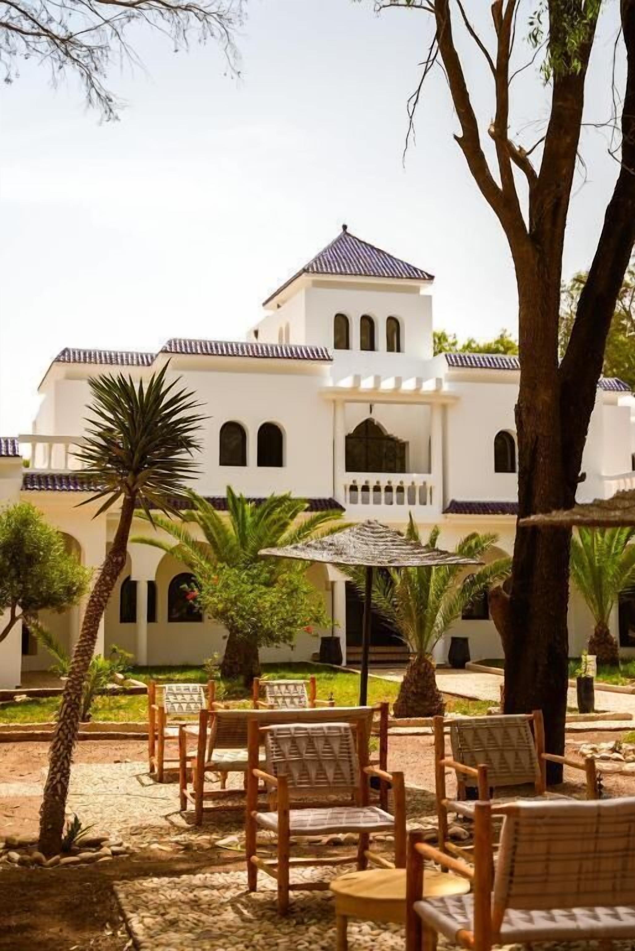 A building with a white facade, arched windows, and a central tower. There are outdoor seating areas with chairs and tables.