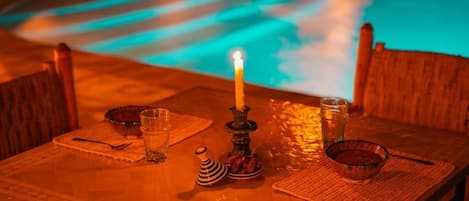 A table with a candle, glasses, and a bowl, set against a backdrop of a pool with a blue hue.
