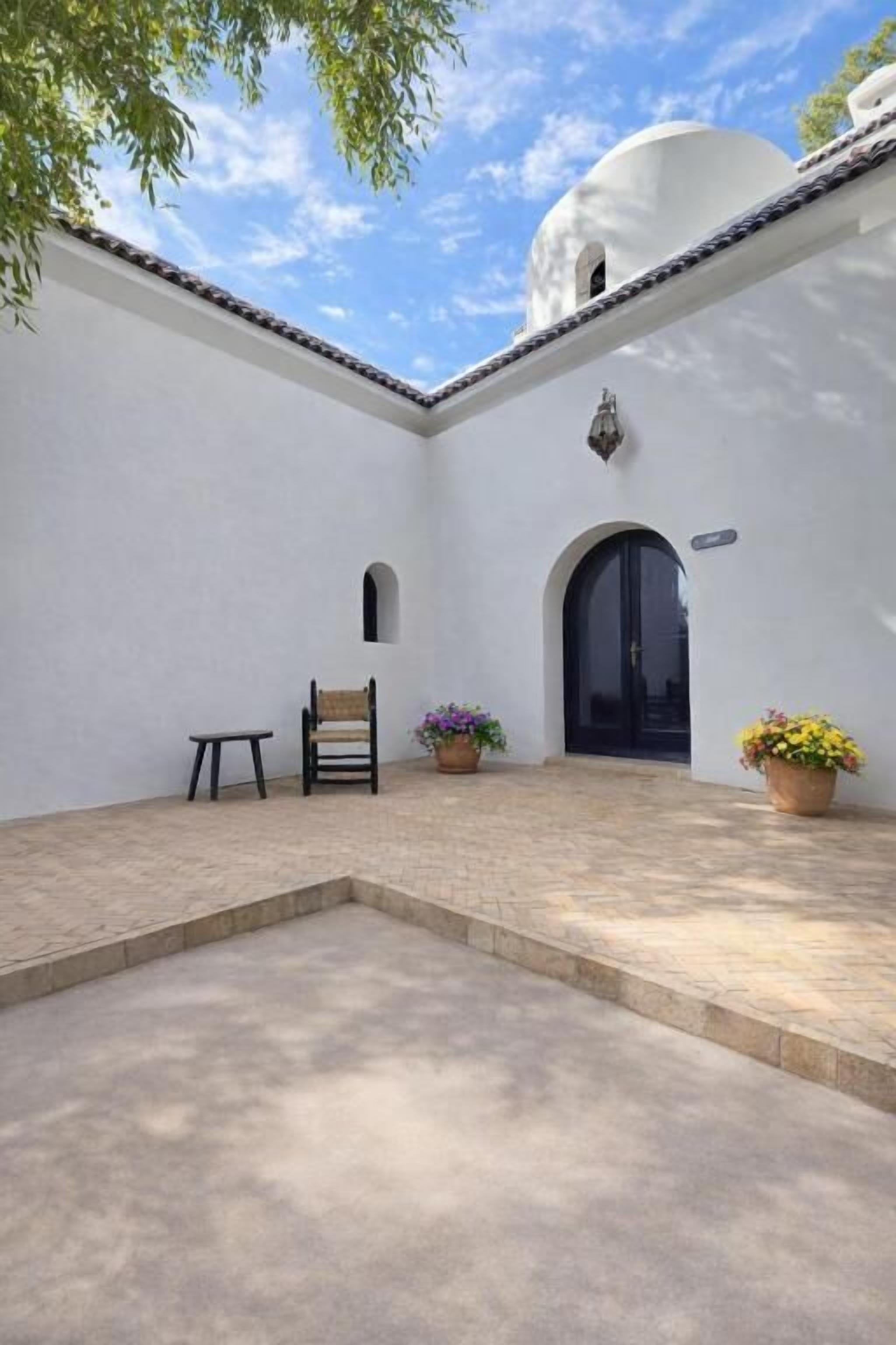 Un patio con un edificio blanco, una puerta negra, un banco y plantas en macetas.