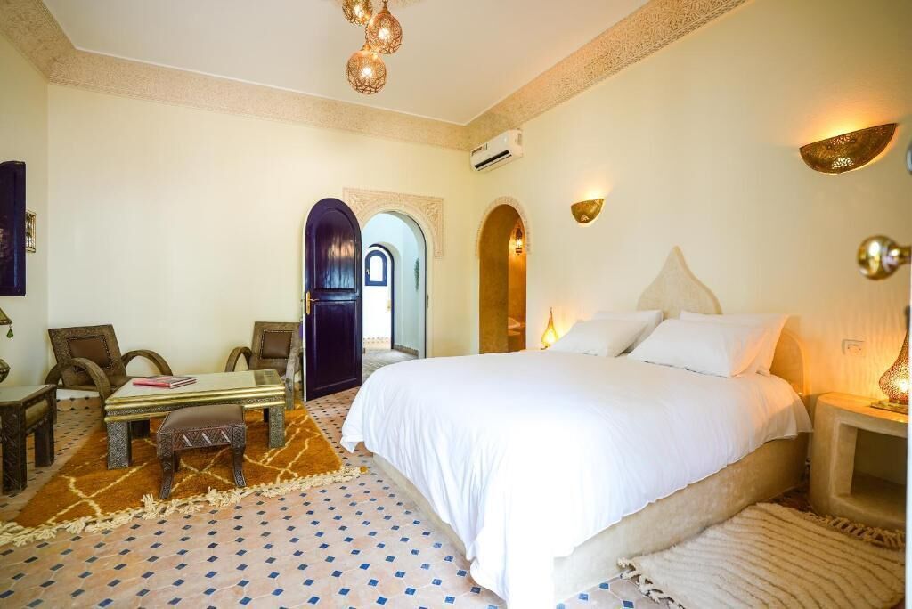 A hotel room with a bed, a desk, chairs, and a patterned floor.