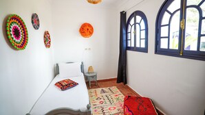 A room with a bed, a small table, a lamp, a patterned rug, and decorative wall hangings.