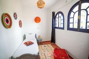 A room with a bed, a small table, a lamp, a patterned rug, and decorative wall hangings.