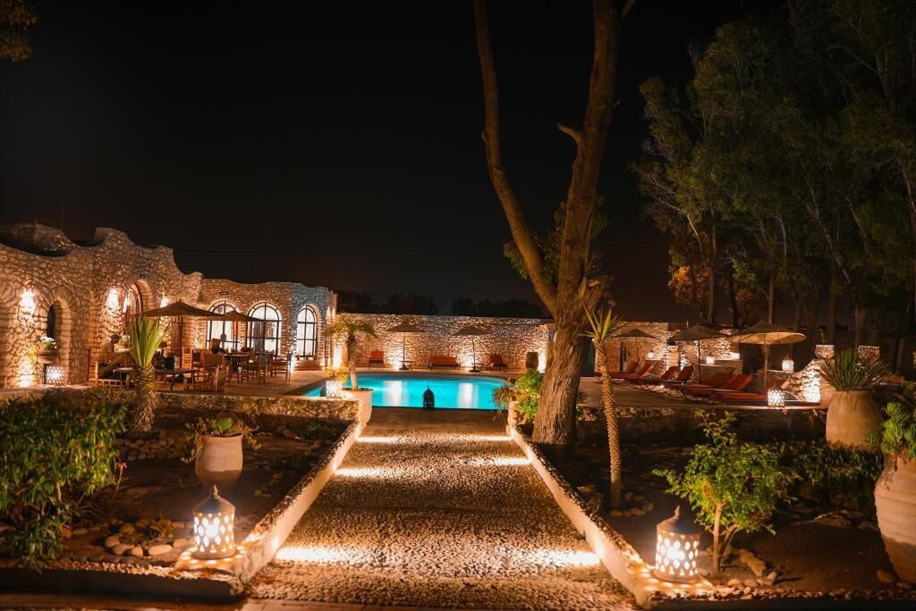 Vue nocturne d’un complexe hôtelier doté d’une piscine, d’un espace de détente en plein air et d’une architecture en pierre.