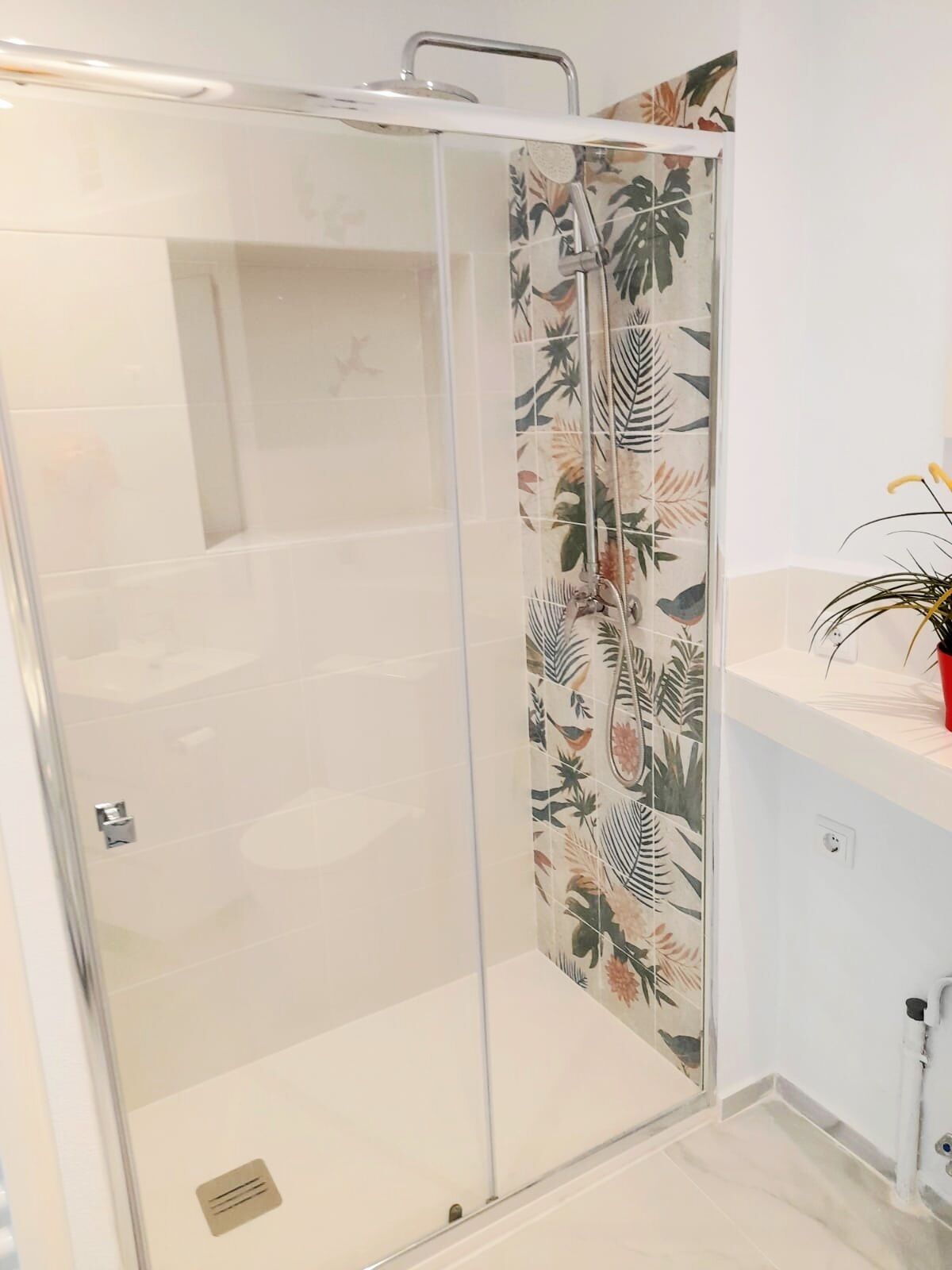 A glass-enclosed shower with a floral tile design, a white sink with a plant, and a white tiled floor.