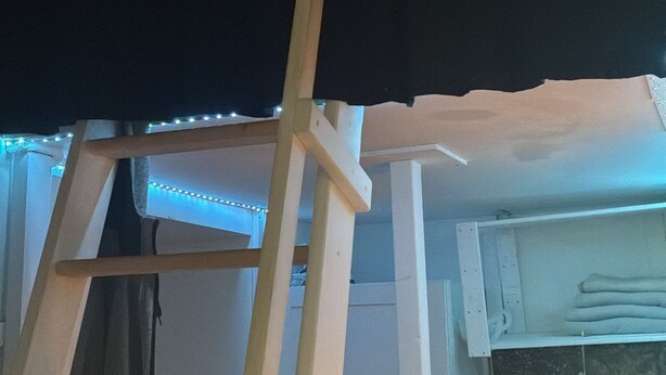 Een kamer met een ladder tegen een muur, een zwart gordijn en een plank met opgevouwen kleding en een dromenvanger.