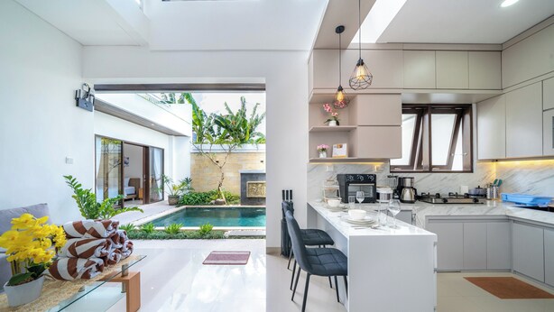 A modern kitchen with a central island, a dining area with chairs, and a pool area with a view of the outdoors. - Addikha Villa by Betterplace (Canggu)