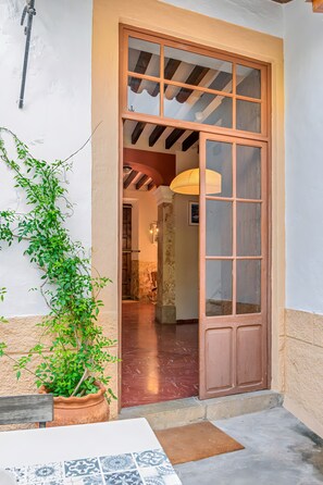 Una puerta con marco de madera y paneles de vidrio, que da acceso a un interior con piso de baldosas y una lámpara colgante.