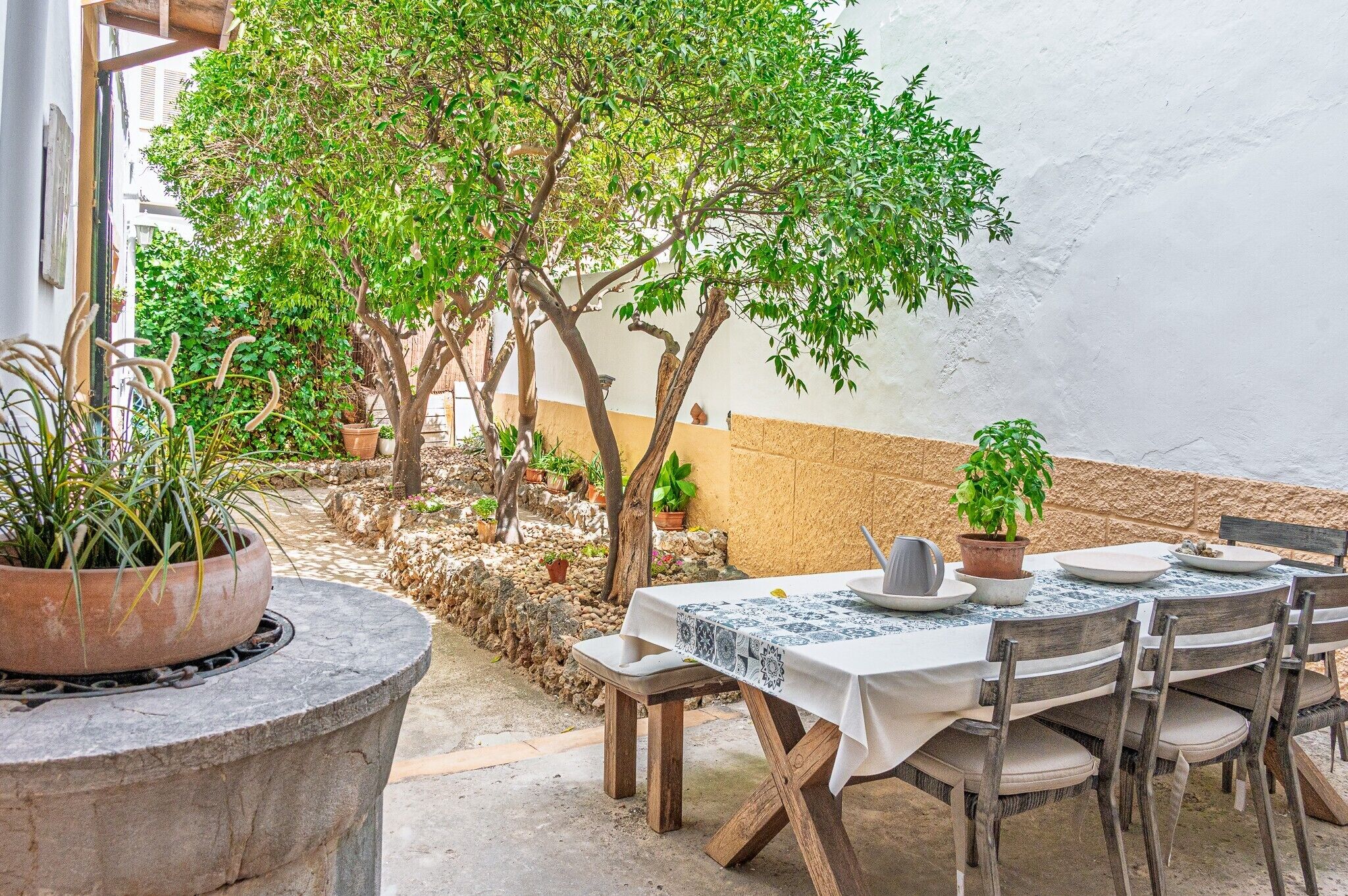 An outdoor dining area with a table set for two, surrounded by potted plants and a tree.