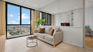 A modern living room with a sofa, a coffee table, and a large window offering a city view. - Brunn by Kozystay - Cilandak (South Jakarta)