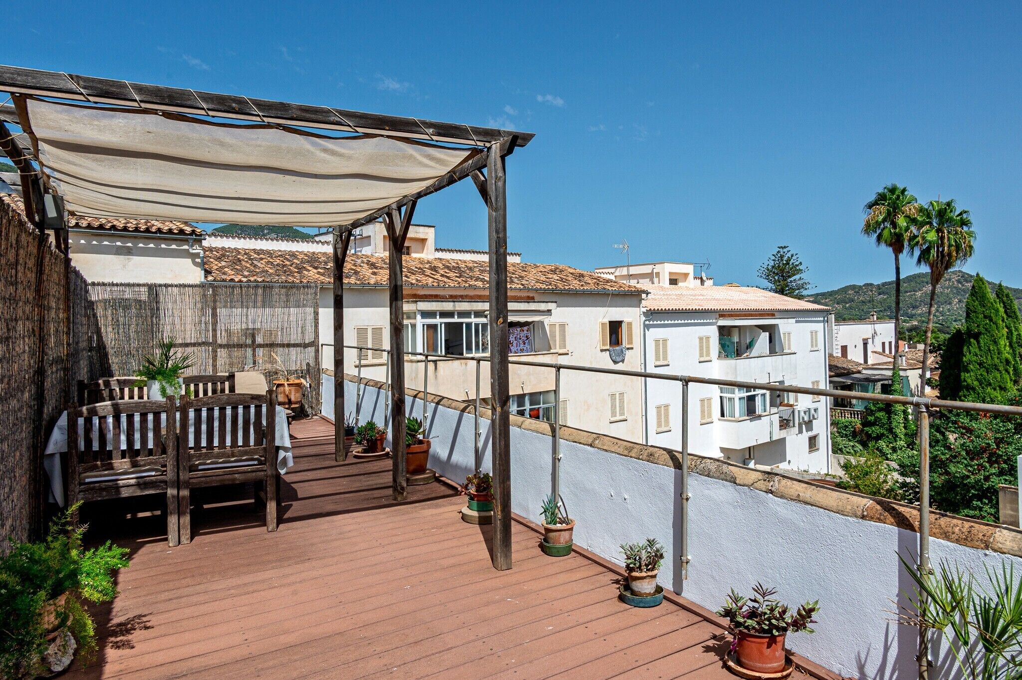 En träveranda med en bänk och krukväxter, med utsikt över ett bostadsområde med vita byggnader och en klar himmel.