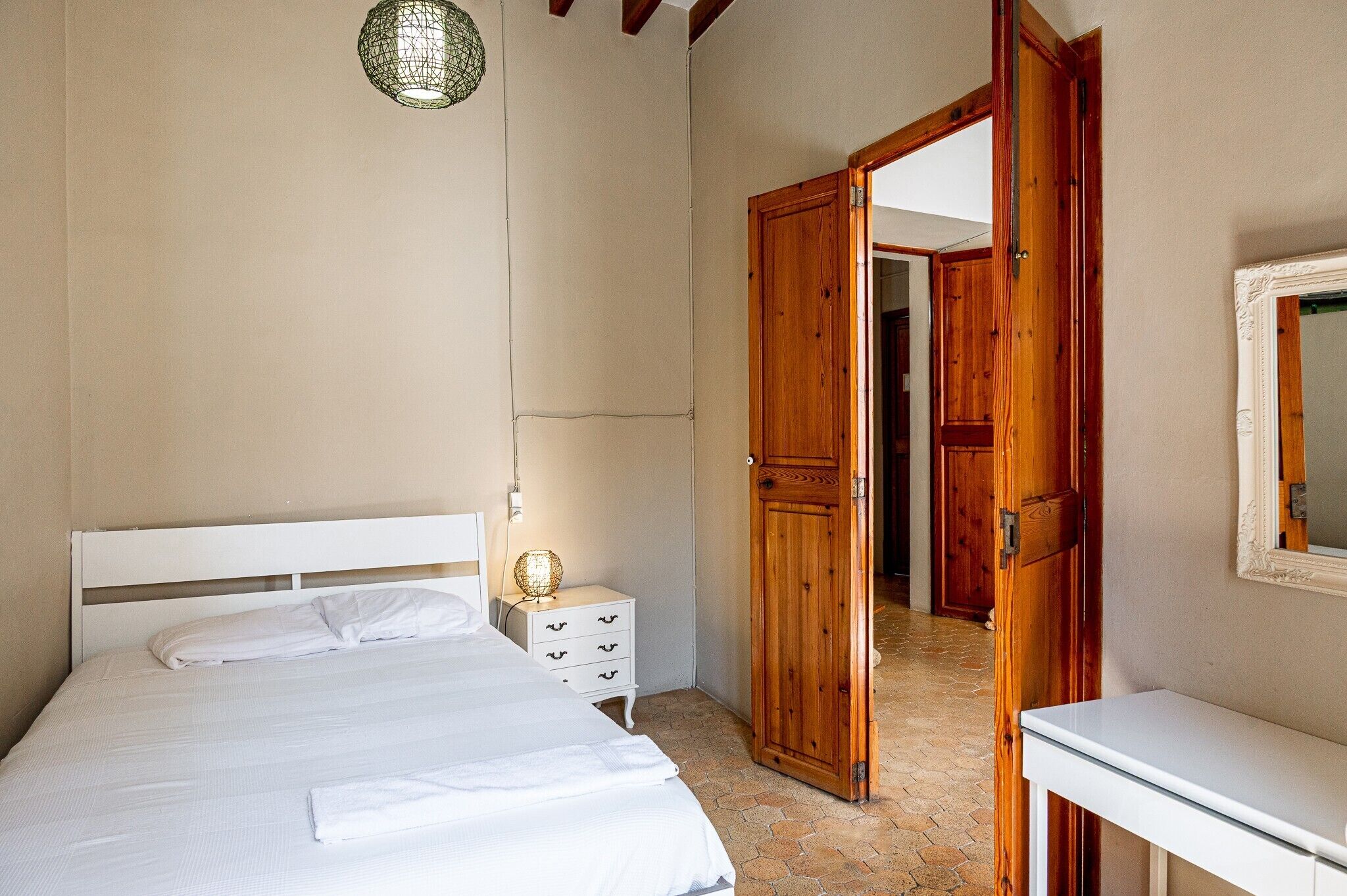 Un dormitorio con cama, mesita de noche, lámpara y una puerta de madera abierta que da a otra habitación.