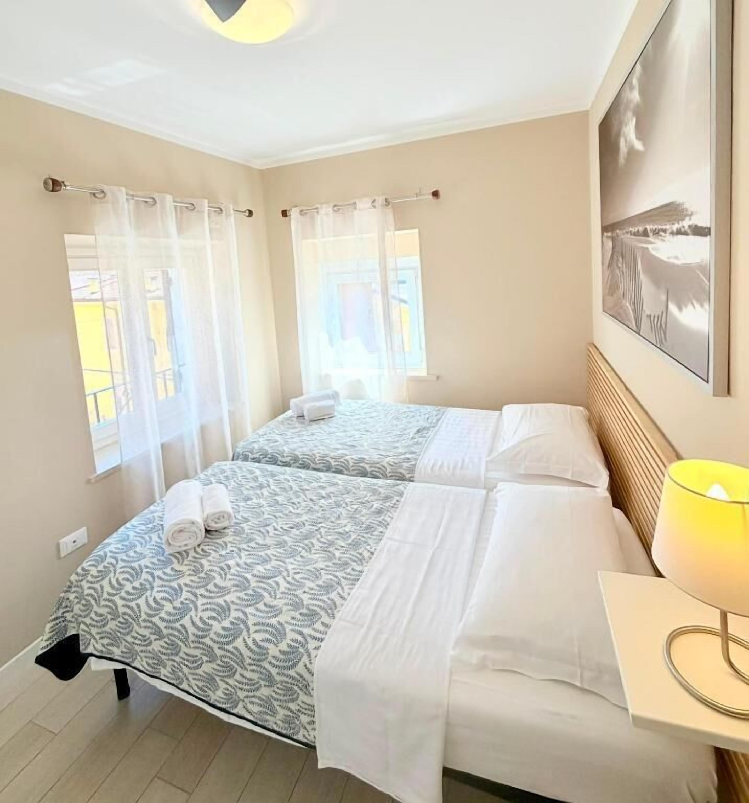 A hotel room with two beds, a bedside table with a lamp, and a window with curtains.
