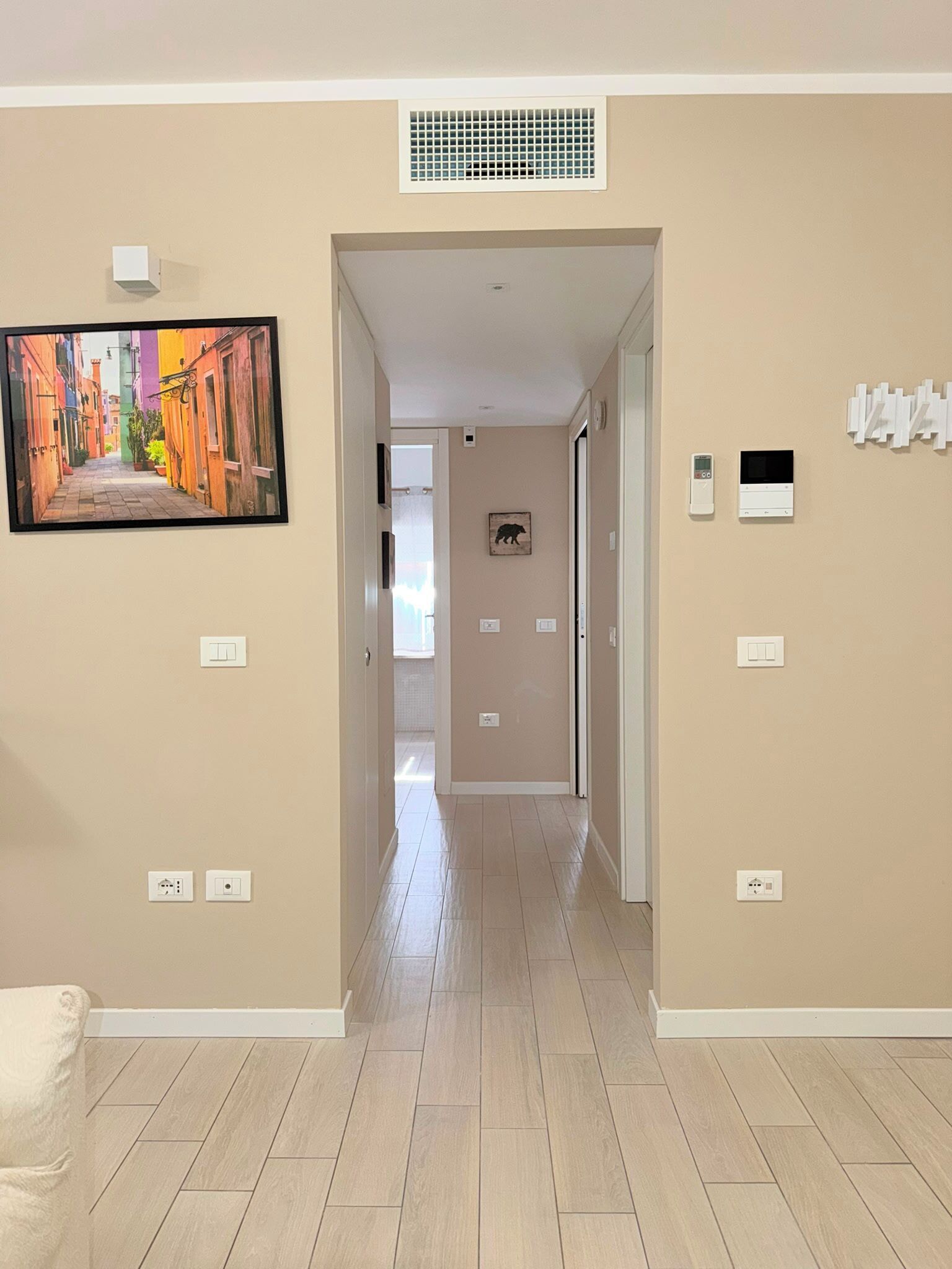 A hallway with beige walls, a tiled floor, and a painting of a colorful street scene. There are light switches and a thermostat on the walls.