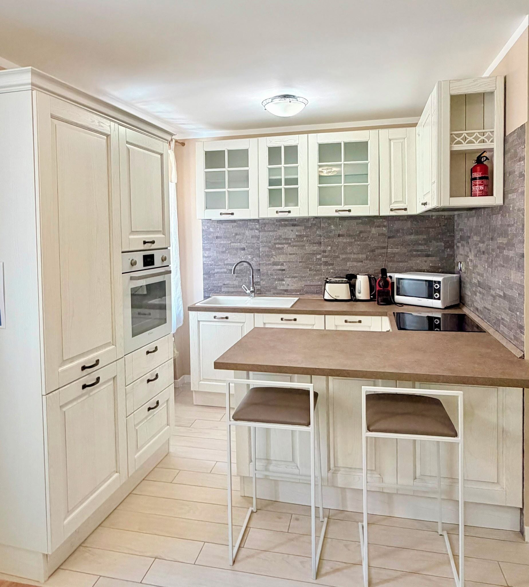 A kitchen with white cabinetry, a tiled backsplash, a double oven, a sink, and two bar stools.