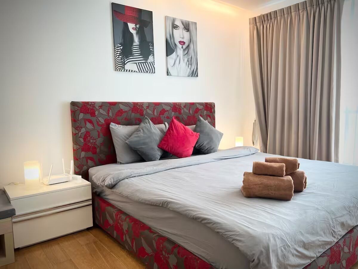 A neatly made bed with a grey and red floral headboard, a bedside table with a lamp, and two wall art pieces above the bed.