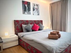 Ein ordentlich bezogenes Bett mit grau-roter Blumenbordwand, ein Nachttisch mit Lampe und zwei Wandbilder über dem Bett.