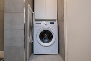 Eine kompakte, weiße Waschmaschine mit Frontlader in einem kleinen, mit Fliesen ausgelegten Waschraum mit einem Wäscheständer.