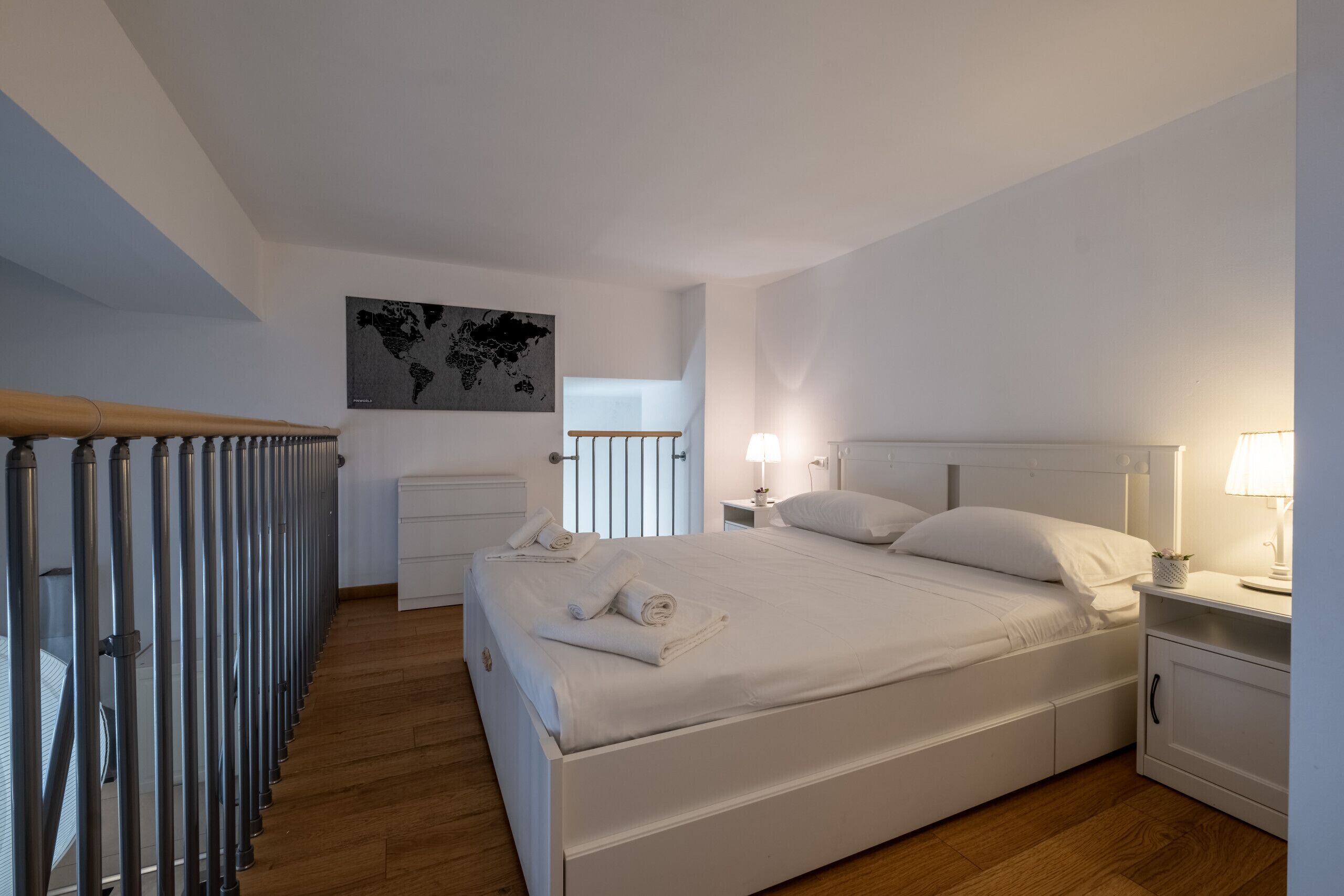 A hotel room with a bed, bedside tables, a nightstand, a lamp, a world map on the wall, and a view of the outside.
