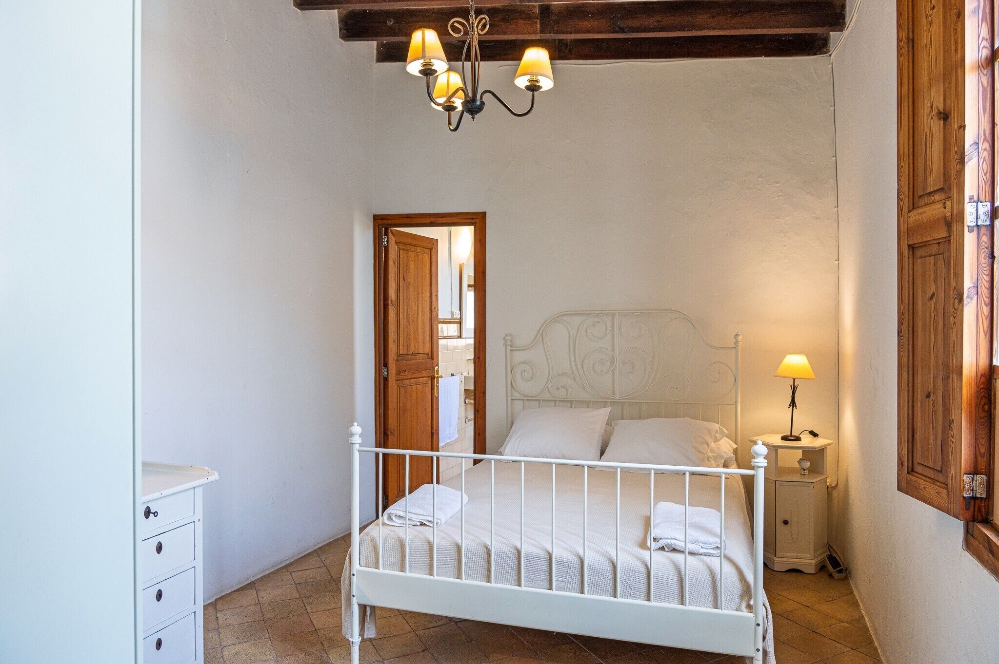 A bedroom with a white metal bed, a wooden door, a white dresser, and a chandelier with three lamps.