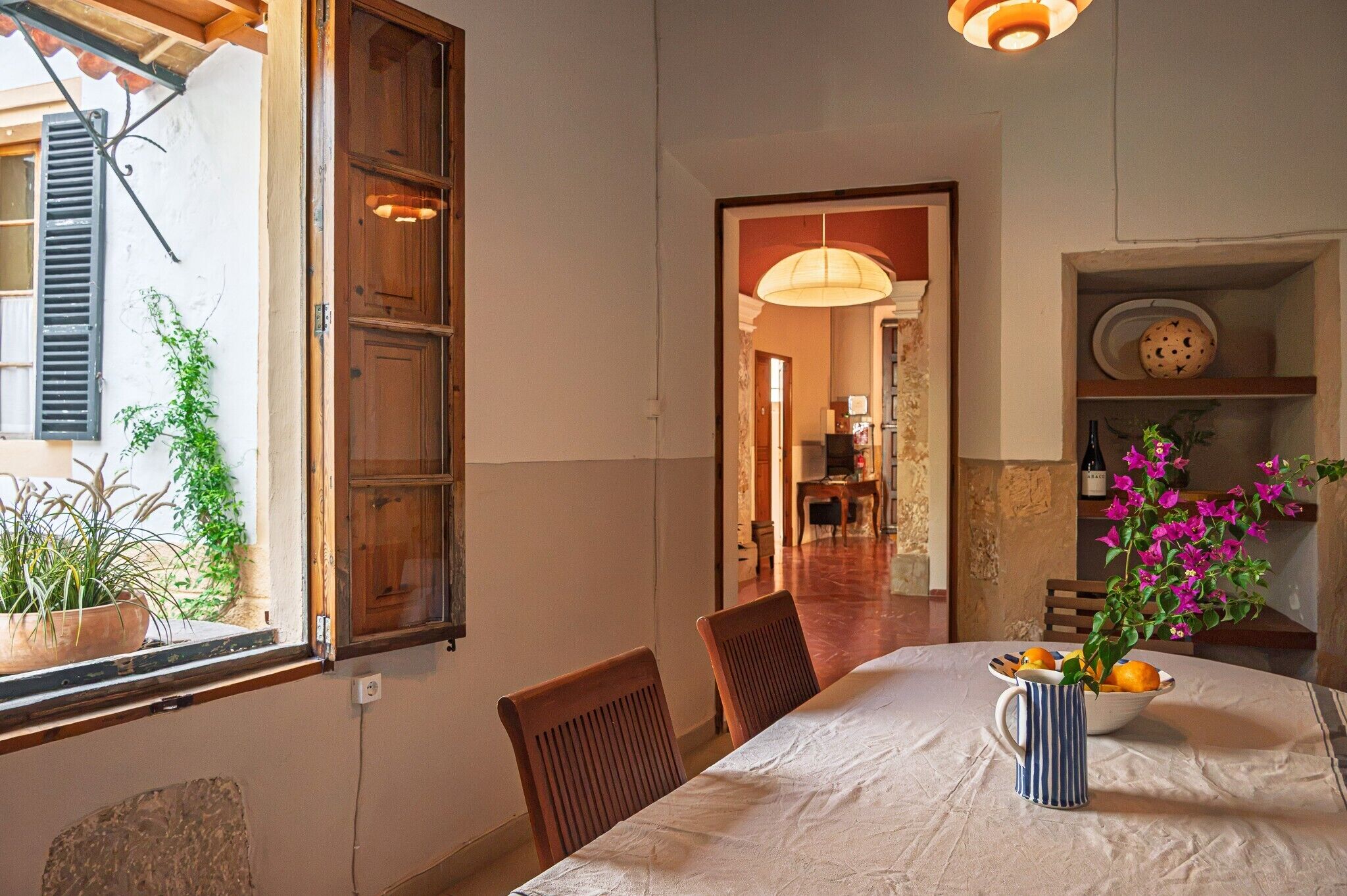 A dining area with a table set for two, chairs, a vase with flowers, and a window with shutters.