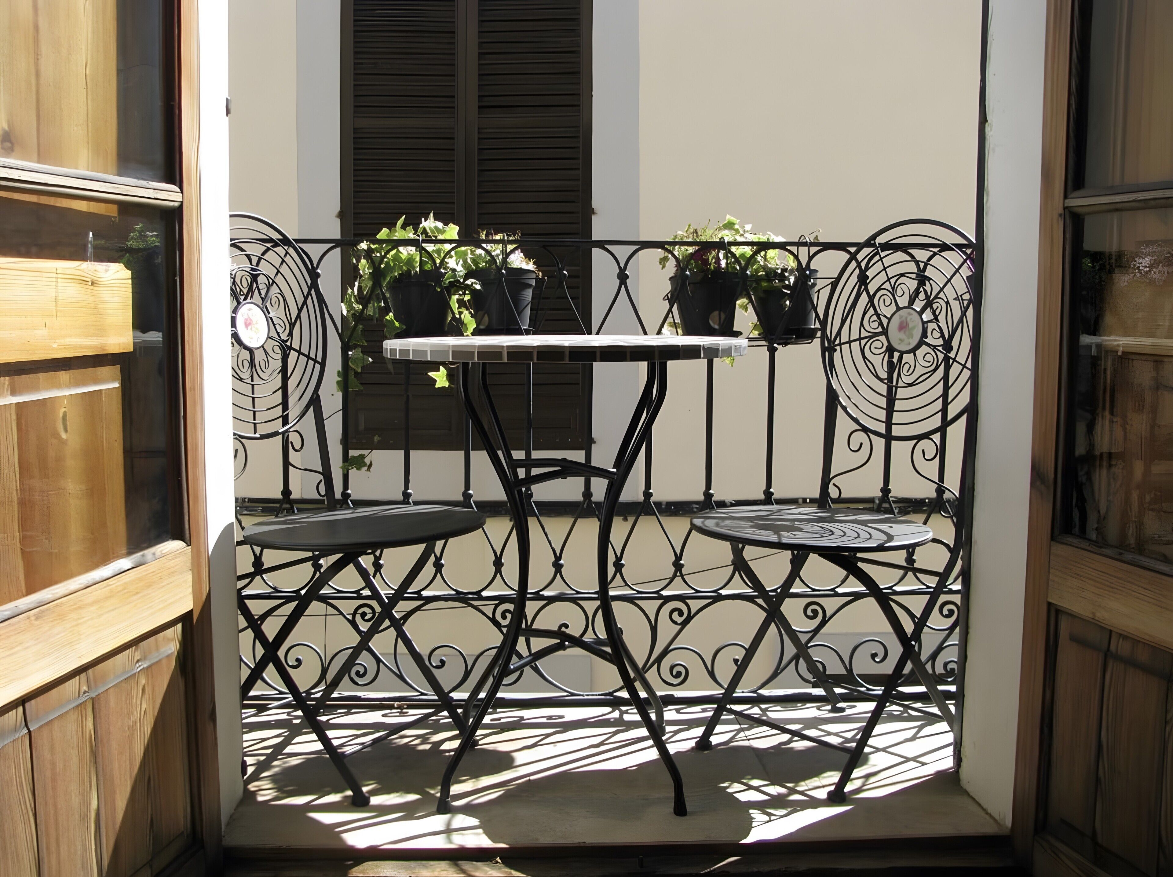 A small outdoor seating area with a table and two chairs, surrounded by a decorative metal fence, and potted plants on the fence.