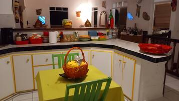 Une cuisine avec une table jaune et des chaises vertes, un panier de fruits et divers ustensiles de cuisine sur le plan de travail.