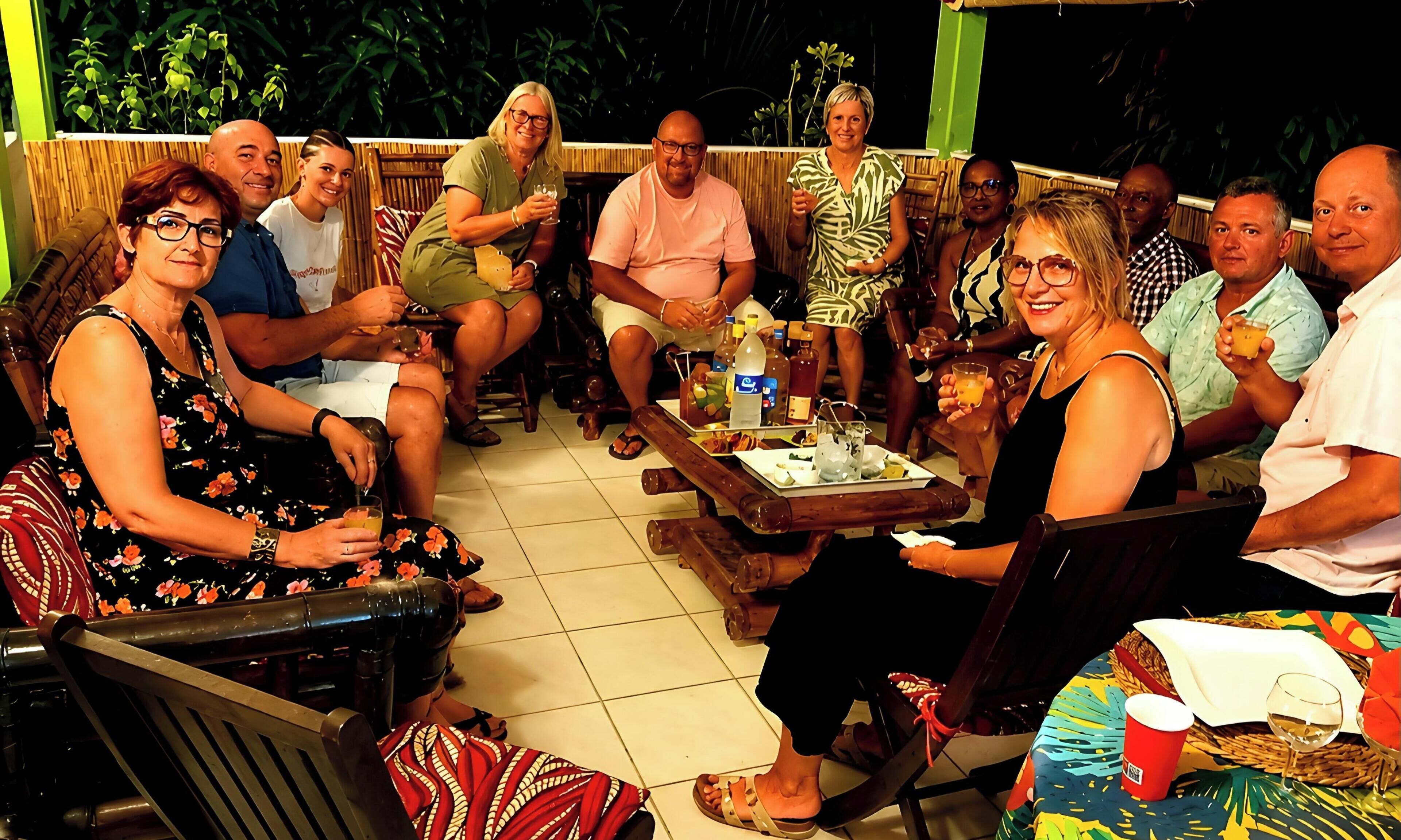 A group of people seated around a wooden table with drinks and snacks, in an outdoor setting with a bamboo fence and tropical plants in the background.