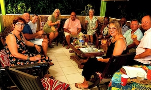 Un groupe de personnes est assis autour d’une table en bois, devant des boissons et des collations, dans un cadre extérieur agrémenté d’une clôture en bambou et de plantes tropicales en arrière-plan.