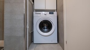 A compact, white front-loading washing machine in a small, tiled laundry room with a metal drying rack. - Caduti di Via Fani by Wonderful Italy (Bologna)