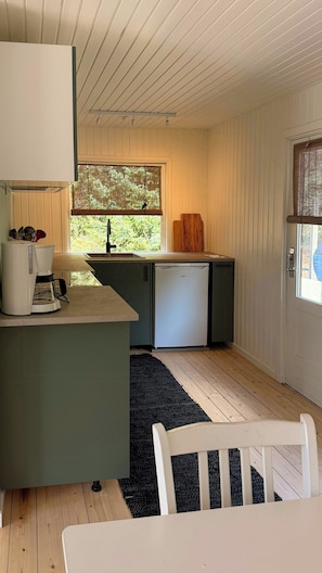 Eine kompakte Küche mit einer Kaffeemaschine, einem Kühlschrank und einem Fenster mit Blick auf Grünflächen.