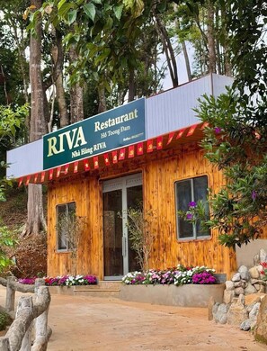 A wooden building with a sign for RIVA Restaurant, surrounded by trees and a flower bed.