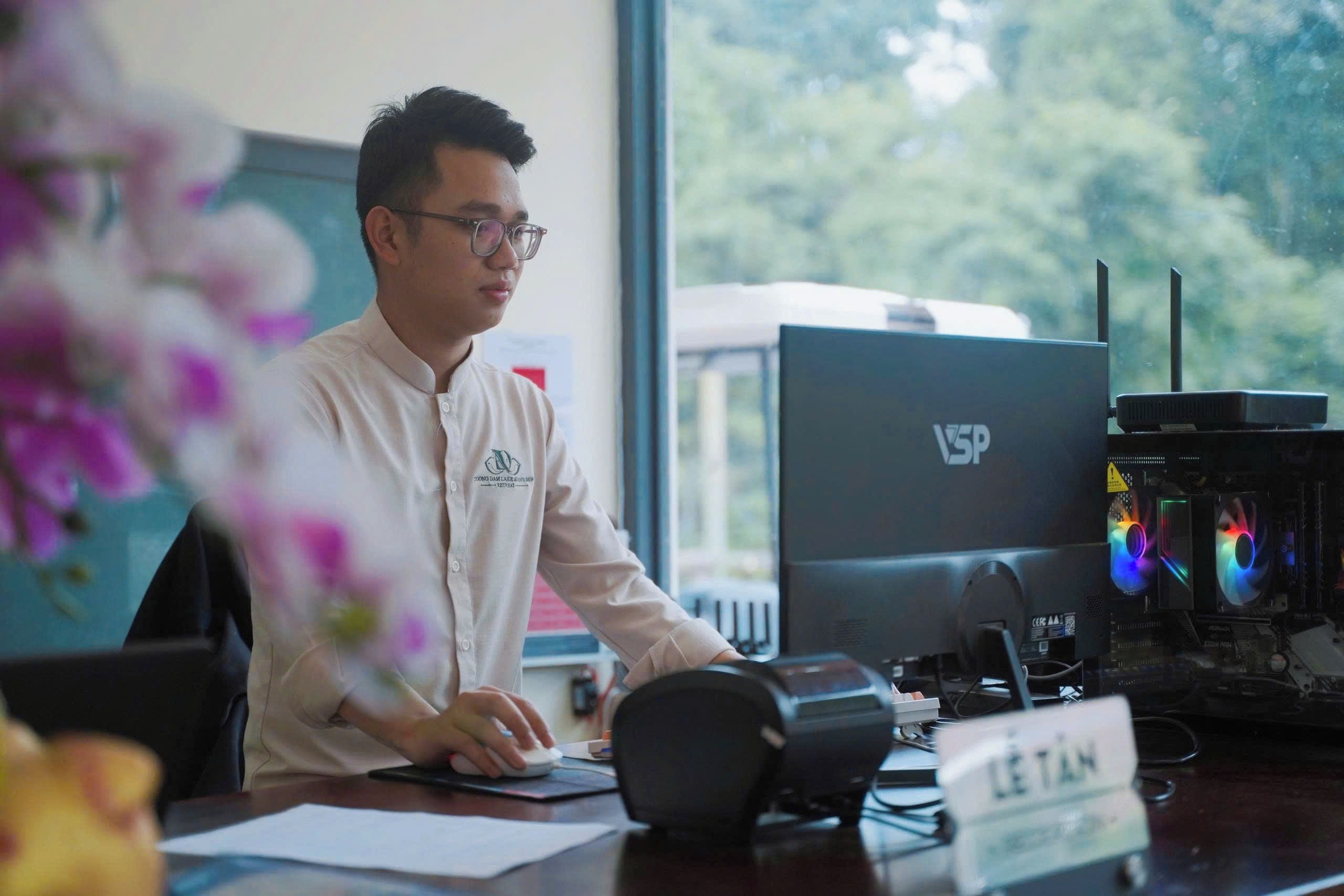 A person seated at a desk with a computer and a monitor, a nameplate, and a window with a view of trees.