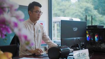 A person seated at a desk with a computer and a monitor, a nameplate, and a window with a view of trees.