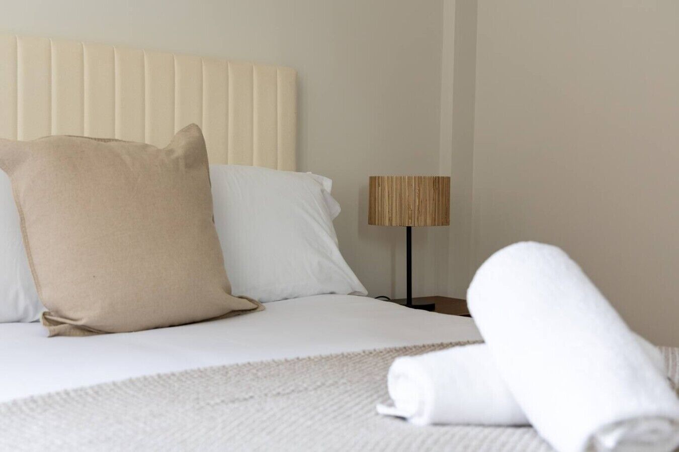 A neatly made bed with white linens and a beige pillow, a wooden floor lamp, and a rolled white towel on the bed.