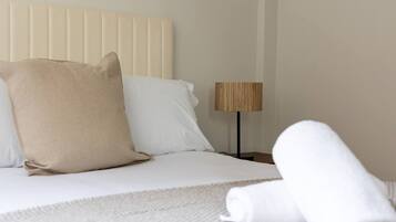 A neatly made bed with white linens and a beige pillow, a wooden floor lamp, and a rolled white towel on the bed.