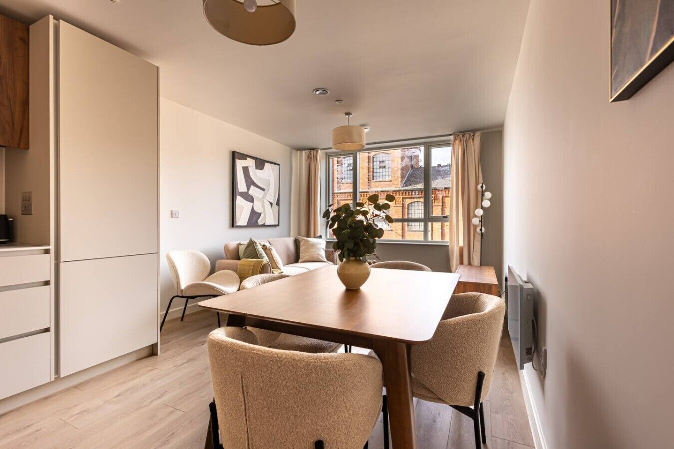 A modern kitchen with a dining area, a sofa set, and a large window with a view of buildings.
