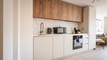 A modern kitchen with white cabinets, a wooden upper shelf, a microwave, a kettle, and a stove.