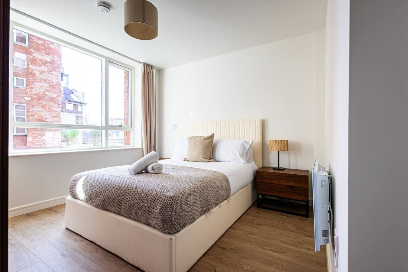 A modern bedroom with a large bed, a bedside table, a lamp, and a window with a view of buildings.