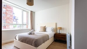 Moderni makuuhuone, jossa on suuri sänky, yöpöytä, lamppu ja ikkuna, josta on näkymä rakennuksiin.