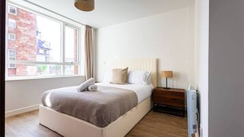 A modern bedroom with a large bed, a bedside table, a lamp, and a window with a view of buildings.