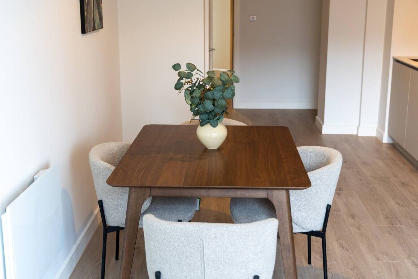 A dining area with a wooden table and two chairs, a vase with green foliage, and a hallway in the background.
