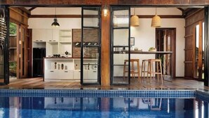 A modern kitchen with glass doors, a dining area with stools, and a pool in the foreground.